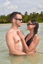 Happy couple in sunglasses embracing in shallow tropical water off a palm-lined beach, woman in a black bikini and shirtless man smiling and gazing at each other.