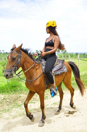 Woman wearing a yellow helmet and black activewear horseback riding a brown horse along a dirt path through green tropical countryside with palm trees and a bright sky