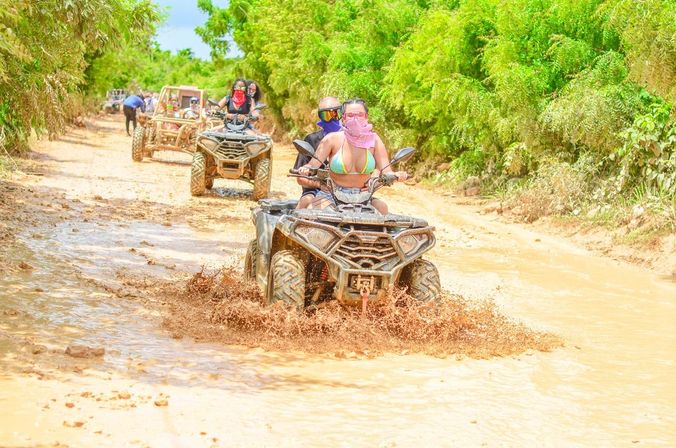 Couple riding an ATV splashing through a muddy tropical dirt trail, followed by other ATVs and a buggy on a sunny off‑road adventure