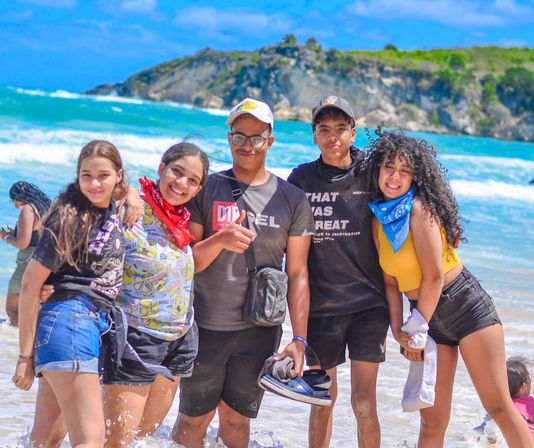 Five smiling teens in casual summer clothes and colorful bandanas posing on a sunny turquoise beach with waves and a rocky headland in the background