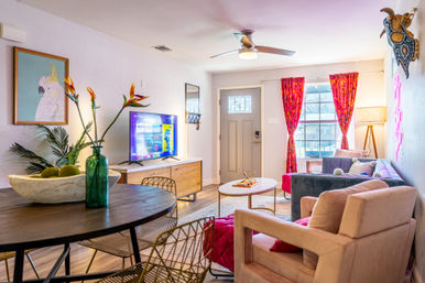 Bright, sunlit modern living room with pink and gray seating, hardwood floors, TV on a wooden console, round table with green vase and bird-of-paradise flowers, patterned pink curtains and front entry door.