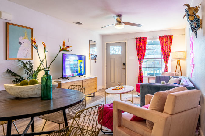 Bright, sunlit modern living room with pink and gray seating, hardwood floors, TV on a wooden console, round table with green vase and bird-of-paradise flowers, patterned pink curtains and front entry door.