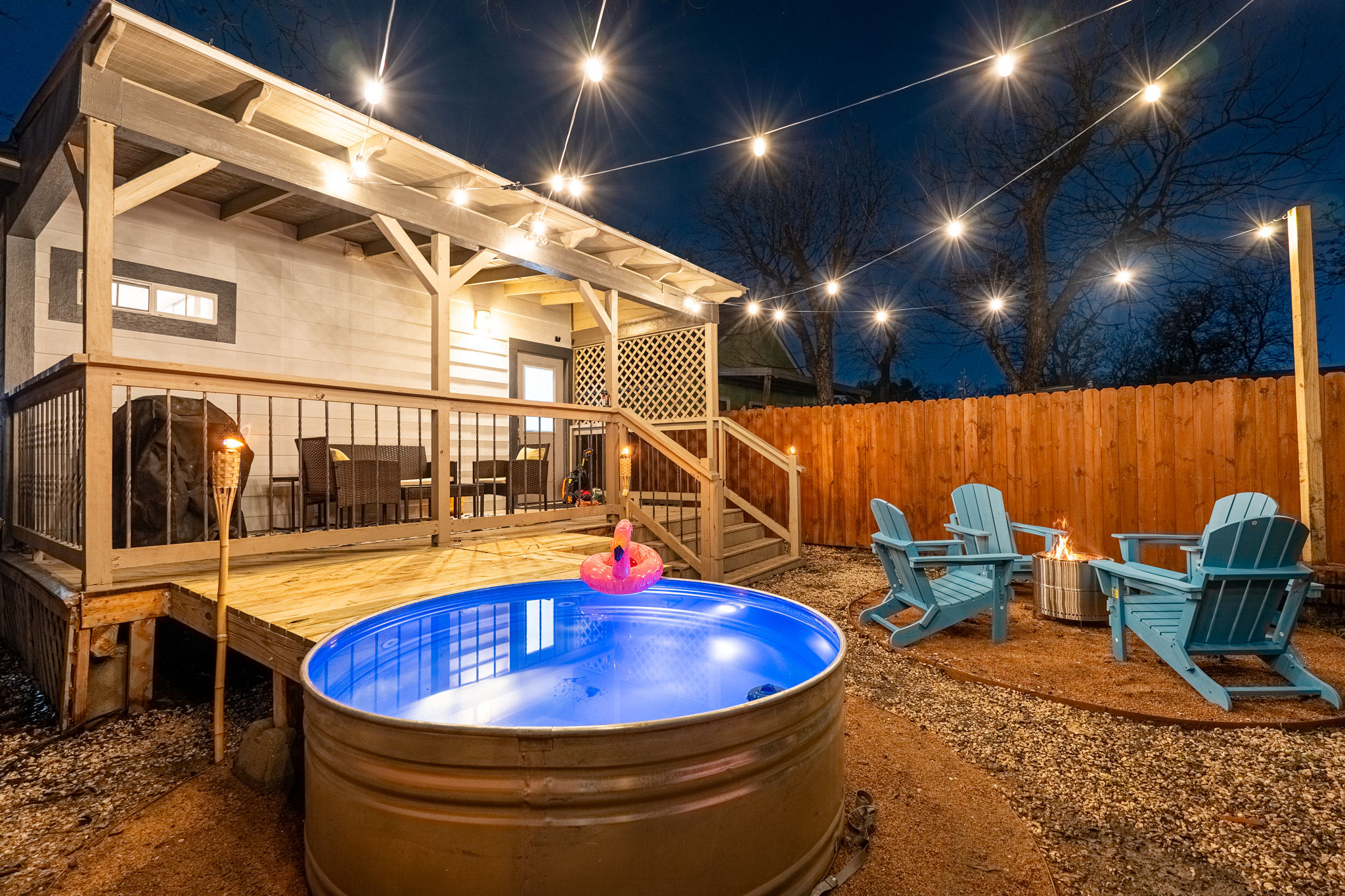 Cozy backyard patio at night with string lights, covered wooden deck, glowing blue metal stock-tank pool with inflatable flamingo, Adirondack chairs around a fire pit and wooden privacy fence