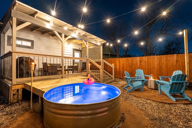 Cozy backyard patio at night with string lights, covered wooden deck, glowing blue metal stock-tank pool with inflatable flamingo, Adirondack chairs around a fire pit and wooden privacy fence