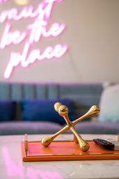Gold decorative jack on an orange tray with a TV remote on a marble coffee table in a modern living room with a teal sofa and pink neon wall sign blurred in the background