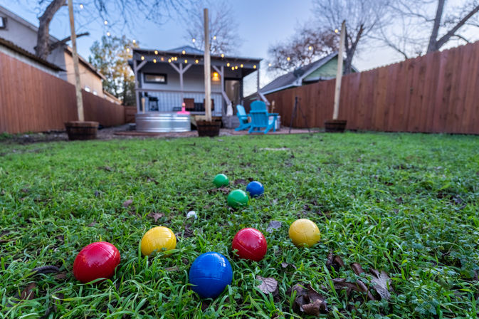 Playful colorful bocce balls scattered on a green backyard lawn in front of a small deck with blue chairs, metal tub, wooden fence, and string lights.
