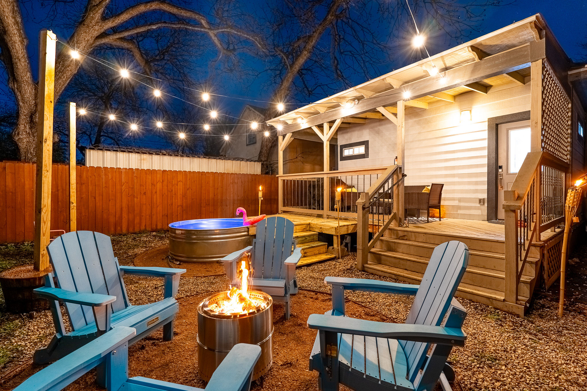 Cozy backyard patio at dusk with string lights over a wooden deck and covered porch, blue Adirondack chairs around a metal fire pit, and a round hot tub with a pink flamingo float.