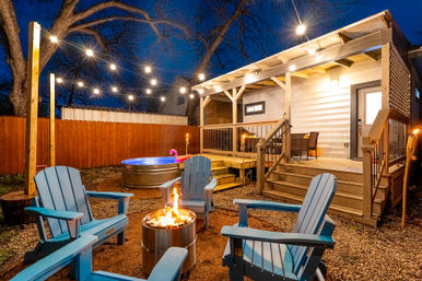 Cozy backyard patio at dusk with string lights over a wooden deck and covered porch, blue Adirondack chairs around a metal fire pit, and a round hot tub with a pink flamingo float.