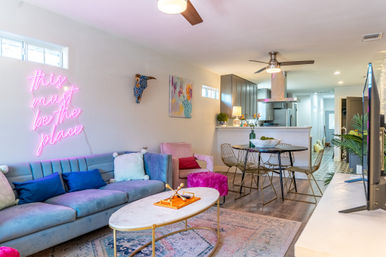 Bright, playful open-plan living room and kitchen in a modern apartment — pastel velvet sofa with colorful pillows, neon pink wall sign reading “this must be the place,” marble coffee table, round dining table with wire chairs, ceiling fan and stainless-steel range hood.