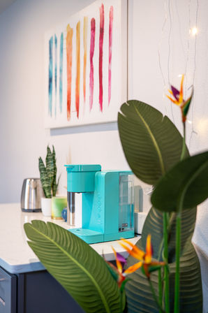 Turquoise single-serve coffee maker on a white kitchen counter by a stainless kettle and potted plants, framed colorful stripe artwork on the wall and bird-of-paradise leaves in the foreground — modern home coffee station.
