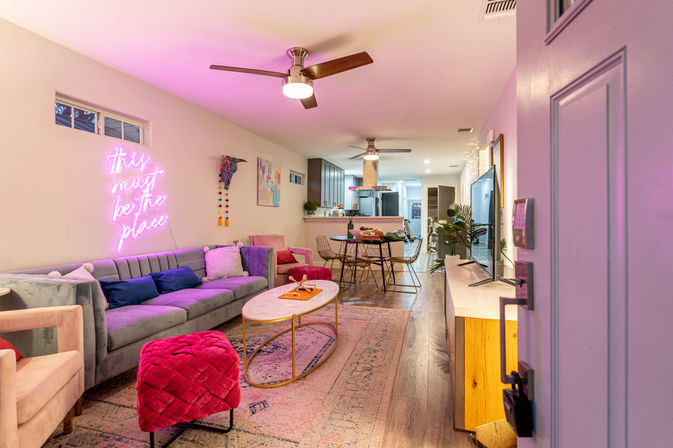 Modern open-plan apartment living room and kitchen with pink neon sign reading 'this must be the place', gray velvet sofa, fuchsia ottoman, marble coffee table, dining table, wood floors and ceiling fans with warm pink accent lighting.