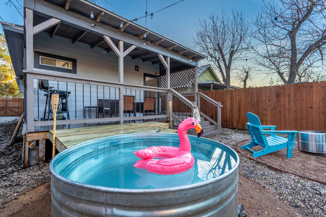 Cozy suburban backyard with a metal stock-tank pool holding a pink flamingo float, covered wooden porch with patio seating and grill, blue Adirondack chair and fenced yard at dusk