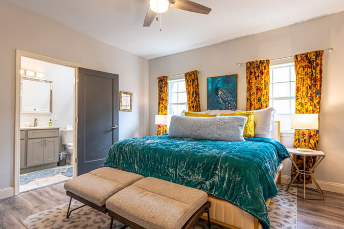 Cozy bright bedroom with a teal velvet comforter and plush pillows on a king bed, peacock wall art between two windows with yellow floral curtains, bedside lamps, two ottomans at the foot of the bed, ceiling fan overhead and an open door revealing an ensuite bathroom.
