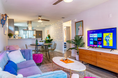 Bright, cozy open-plan apartment living room with pastel velvet sofa, pink ottoman, marble coffee table, wooden TV console showing streaming apps, round dining table and open kitchen, string fairy lights and houseplants.