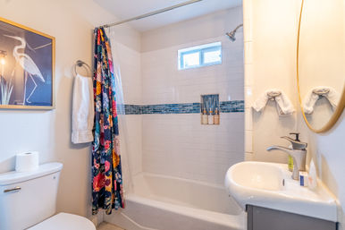 Bright modern bathroom with white bathtub and subway-tile shower, blue glass mosaic accent strip, cheerful floral shower curtain, small window, pedestal sink and toilet.