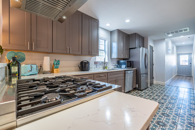 Bright modern galley kitchen with stainless gas cooktop, gray shaker cabinets, quartz countertops, stainless refrigerator and playful blue patterned tile floor.