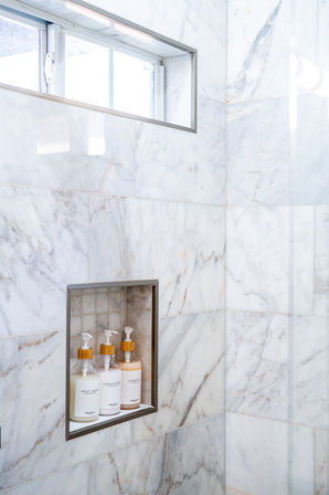 Sleek white marble shower with a recessed niche holding three matching pump bottles labeled body wash, conditioner and shampoo beneath a narrow window — modern bathroom detail.