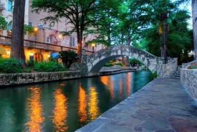 Stone arched footbridge over a calm canal at dusk, warm lantern reflections rippling on the water, tree-lined riverside walkway and stone buildings.