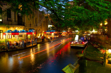 Vibrant nighttime riverwalk scene: lighted tour boat and colorful light trails on the canal, outdoor riverside restaurants under red-and-blue umbrellas, trees and an arched pedestrian bridge.