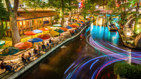 Nighttime urban riverwalk with colorful umbrellas and canal-side outdoor dining under trees, illuminated bridges and vibrant multicolored boat light trails streaking across the water.