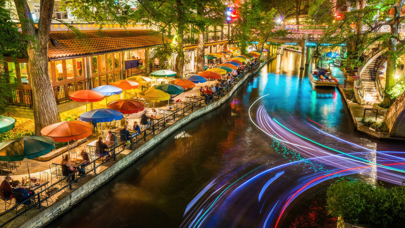 Nighttime urban riverwalk with colorful umbrellas and canal-side outdoor dining under trees, illuminated bridges and vibrant multicolored boat light trails streaking across the water.