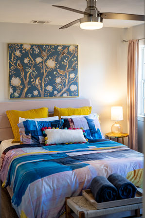 Cozy modern bedroom with vibrant blue-and-yellow bedding, playful pom-pom lumbar pillow, blush headboard, framed bird-and-flower artwork, ceiling fan, glowing bedside lamp, and rolled towels on a bench.