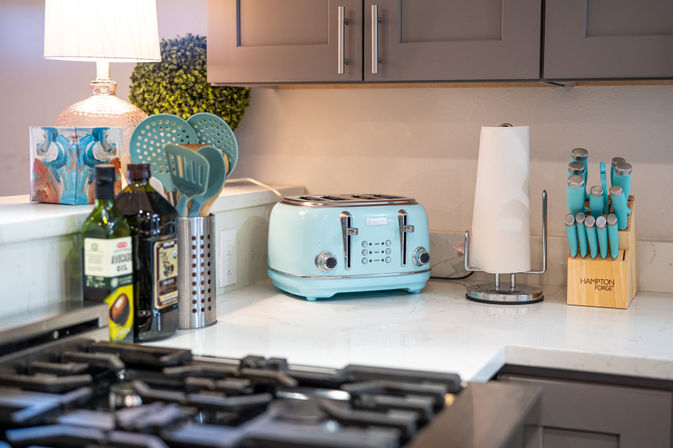 Sunlit modern kitchen countertop with a retro aqua four-slice toaster, matching turquoise knife set in a wooden block, utensil caddy, olive oil bottles, paper towel holder and gas stove in the foreground