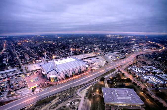 Aerial dusk view of a large lit sports stadium beside a winding highway, surrounded by urban neighborhoods and glowing city lights under a cloudy sky.