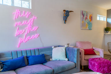 Cozy modern living room decor with pastel velvet sofa and pink armchair, pink neon wall sign reading 'this must be the place', colorful throw pillows, abstract wall art and decorative painted bull skull.