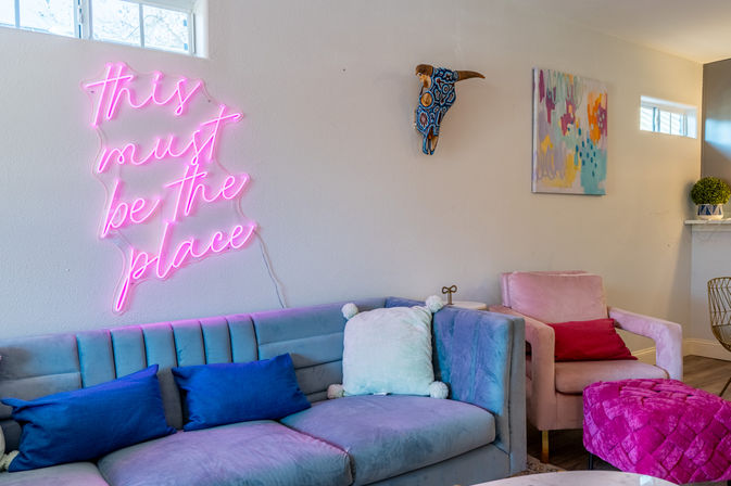 Cozy modern living room decor with pastel velvet sofa and pink armchair, pink neon wall sign reading 'this must be the place', colorful throw pillows, abstract wall art and decorative painted bull skull.