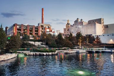 Sunset urban waterfront park with a reflective pond, stone terraces and docks, lit trees and walkways, and historic brick industrial buildings with a tall smokestack on the skyline.