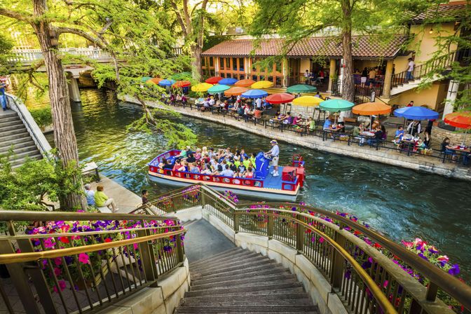 Colorful riverwalk scene with a red tour boat of passengers cruising a tree-lined canal past stone steps and flower-lined railings, with a waterfront promenade of outdoor dining and bright umbrella tables.
