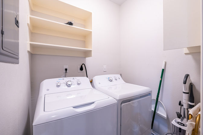 Bright compact laundry room with white top-loading washer and matching dryer, open wall shelves above, and a mop, dustpan and upright vacuum stored beside the machines.