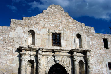 Sunlit stone Spanish colonial mission facade with carved columns, arched entrance and small barred window against a deep blue sky in San Antonio