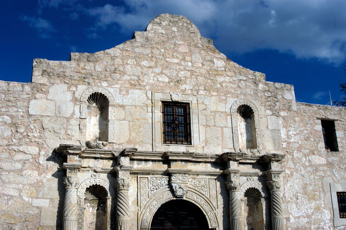 Sunlit stone Spanish colonial mission facade with carved columns, arched entrance and small barred window against a deep blue sky in San Antonio