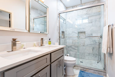 Spa-like modern marble bathroom with double vanity, gray cabinets, dual chrome faucets, glass-enclosed tiled shower, white toilet, towel on rack and blue bath mat.