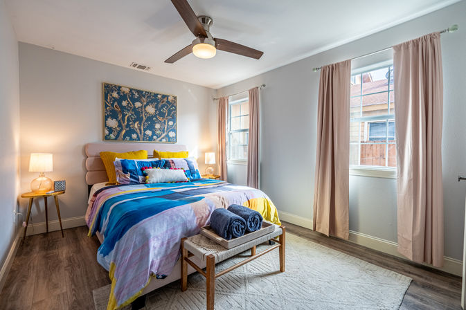 Cozy sunlit bedroom with colorful bedspread, pink upholstered headboard, floral wall art, ceiling fan, two windows with blush curtains, wood floors and a bench with rolled towels.
