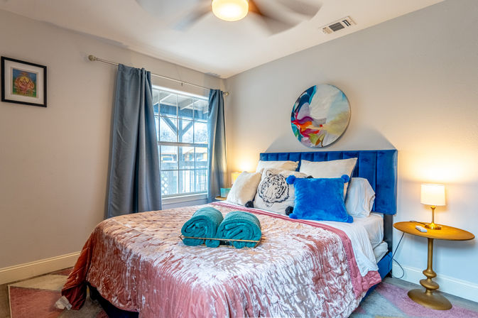 Cozy staged guest bedroom with blue velvet headboard, plush pink velvet bedspread, decorative tiger and blue pillows, teal rolled towels on bed, colorful round wall art, bedside lamps and a window with gray curtains