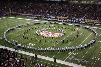 Marching band performing a precision halftime show on a packed indoor football field, band members forming concentric circles around a colorful center logo as fans watch from the stands.