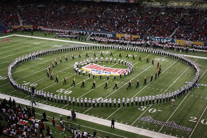 Marching band performing a precision halftime show on a packed indoor football field, band members forming concentric circles around a colorful center logo as fans watch from the stands.