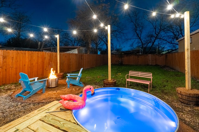 Nighttime backyard scene with glowing blue round plunge pool and pink inflatable flamingo, overhead string lights, lit fire pit with blue Adirondack chairs, wooden fence and grassy yard