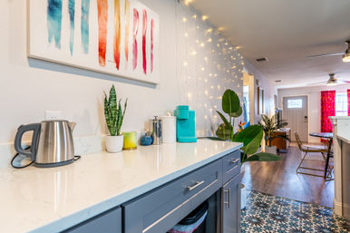 Modern open-plan apartment kitchen with white quartz countertop and blue cabinets, stainless electric kettle and teal coffee maker, potted plants and colorful abstract wall art, string fairy lights on the wall and patterned tile floor transitioning to hardwood toward the dining area and front door