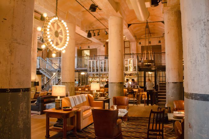 Industrial-chic hotel lobby and bar interior with exposed concrete columns, leather seating, mezzanine level and glowing wheel chandelier