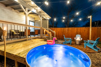 Cozy backyard at dusk featuring a lit stock-tank hot tub with pink flamingo float, wooden deck and covered patio, string lights, fire pit and blue Adirondack chairs by a wooden fence.