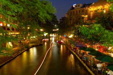 Lively tree-lined riverwalk at night with glowing string lights, riverside restaurants under colorful umbrellas and light-trail boats on a calm canal