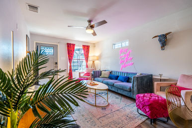 Vibrant boho living room with teal velvet sofa, hot-pink neon sign reading 'this must be the place', bright pink curtains, gold coffee table, patterned rug, leafy plant and decorative bull skull on the wall.