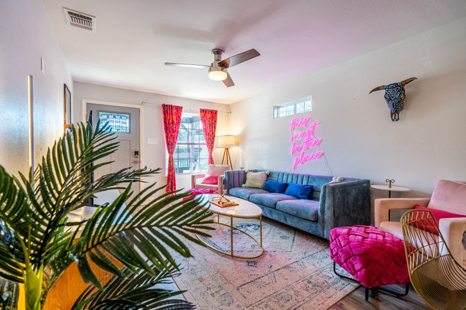 Vibrant boho living room with teal velvet sofa, hot-pink neon sign reading 'this must be the place', bright pink curtains, gold coffee table, patterned rug, leafy plant and decorative bull skull on the wall.