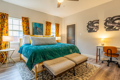 Cozy sunlit bedroom with teal velvet bedspread and fluffy pillows, yellow floral curtains framing two windows, peacock wall art, twin upholstered benches at the foot of the bed, leopard-print rug, desk with an orange chair and warm bedside lamps.