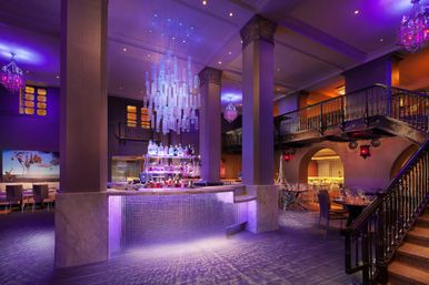 Luxury hotel bar and lounge bathed in moody purple light, featuring a marble-clad illuminated central bar with backlit liquor shelves, a cascading glass chandelier, mezzanine balcony and ornate staircase