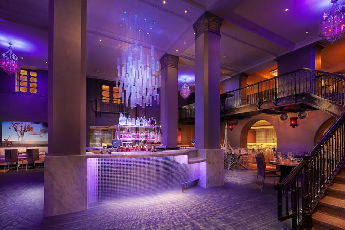 Luxury hotel bar and lounge bathed in moody purple light, featuring a marble-clad illuminated central bar with backlit liquor shelves, a cascading glass chandelier, mezzanine balcony and ornate staircase